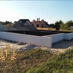 Wylany fundament domu jednorodzinnego z hydroizolacją, widoczne wystające pręty zbrojeniowe, hałdy piasku i w tle dom w trakcie budowy.