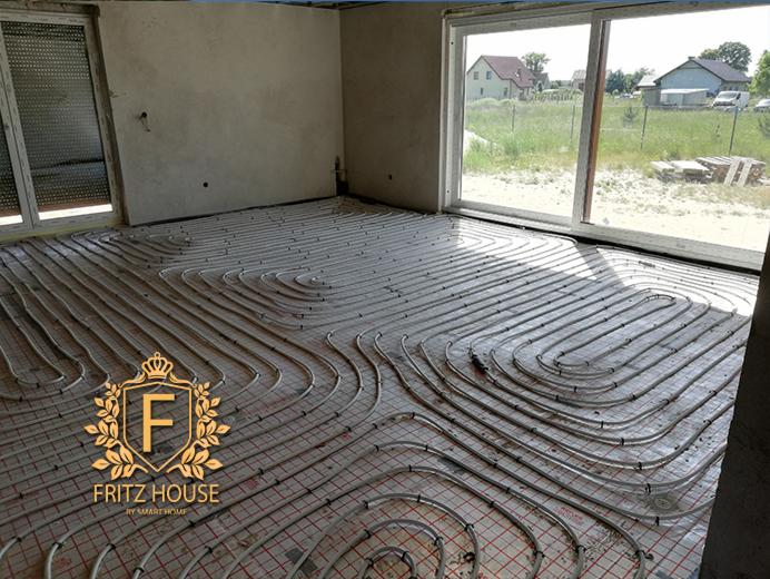 Instalacja ogrzewania podłogowego w surowym wnętrzu domu z widokiem na ogród przez duże okna.