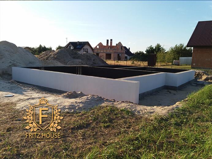 Wylany fundament domu jednorodzinnego z hydroizolacją, widoczne wystające pręty zbrojeniowe, hałdy piasku i w tle dom w trakcie budowy.