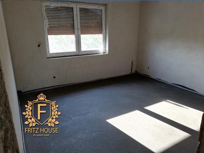 Pokój w surowym stanie z wylewką podłogową, widoczne ślady po położeniu gładzi na ścianach, okno z opuszczonymi roletami, w rogu logo firmy.