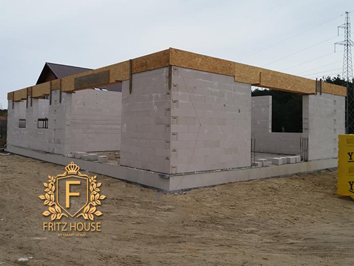 Surowy stan ścian domu z betonu komórkowego z widocznym zbrojeniem i drewnianymi belkami stropowymi, logo firmy Fritz House w lewym dolnym rogu.
