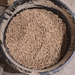 Czarny kubeł wypełniony pelletem drzewnym, widok z góry, obok karton i fragment buta roboczego.