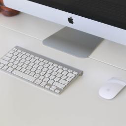 NADERA - firma sprzątająca - Minimalistyczne stanowisko pracy z komputerem iMac, bezprzewodową klawiaturą i myszą na białym biurku, z drewnianym organizerem w tle.