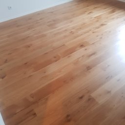 E&P EURO-HOME - Jasnobrązowa podłoga z paneli ułożona w nowym mieszkaniu, widoczne gniazdka elektryczne na białej ścianie w tle.