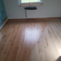 E&P EURO-HOME - Jasny, drewniany parkiet po renowacji, widoczne ślady pyłu i kabel od maszyny szlifującej, okno z widokiem na ogród.