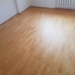 E&P EURO-HOME - Jasnobrązowe panele podłogowe ułożone w pustym pomieszczeniu z białymi ścianami i grzejnikiem żeberkowym.