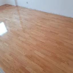 Świeżo ułożona, lakierowana podłoga z paneli imitujących drewno, widoczne odbicie światła, białe ściany w tle.