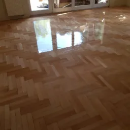 Świeżo wycyklinowany, lakierowany parkiet ułożony w jodełkę, odbijający światło z okna tarasowego.