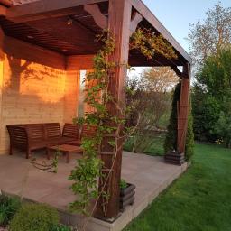 E&P EURO-HOME - Drewniana altana przylegająca do domu z drewnianą boazerią, wyposażona w komplet mebli ogrodowych i pnącą roślinność na słupach konstrukcyjnych, otoczona zielenią ogrodu w ciepłym świetle...