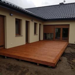 E&P EURO-HOME - Nowoczesny taras z drewna o ciepłym, brązowym odcieniu, przylegający do beżowego domu z ciemnymi oknami i roletami, widok na częściowo przygotowany teren wokół tarasu.