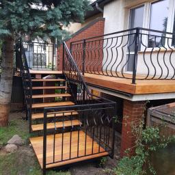 E&P EURO-HOME - Zewnętrzne schody z drewna prowadzące na taras z czarną, dekoracyjną balustradą, przylegające do budynku z ceglanym narożnikiem i dużym oknem.