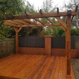 E&P EURO-HOME - Świeżo zaimpregnowany taras drewniany z pergolą w ogrodzie, widoczne krople impregnatu na deskach, w tle mur z cegły i zielona roślinność.