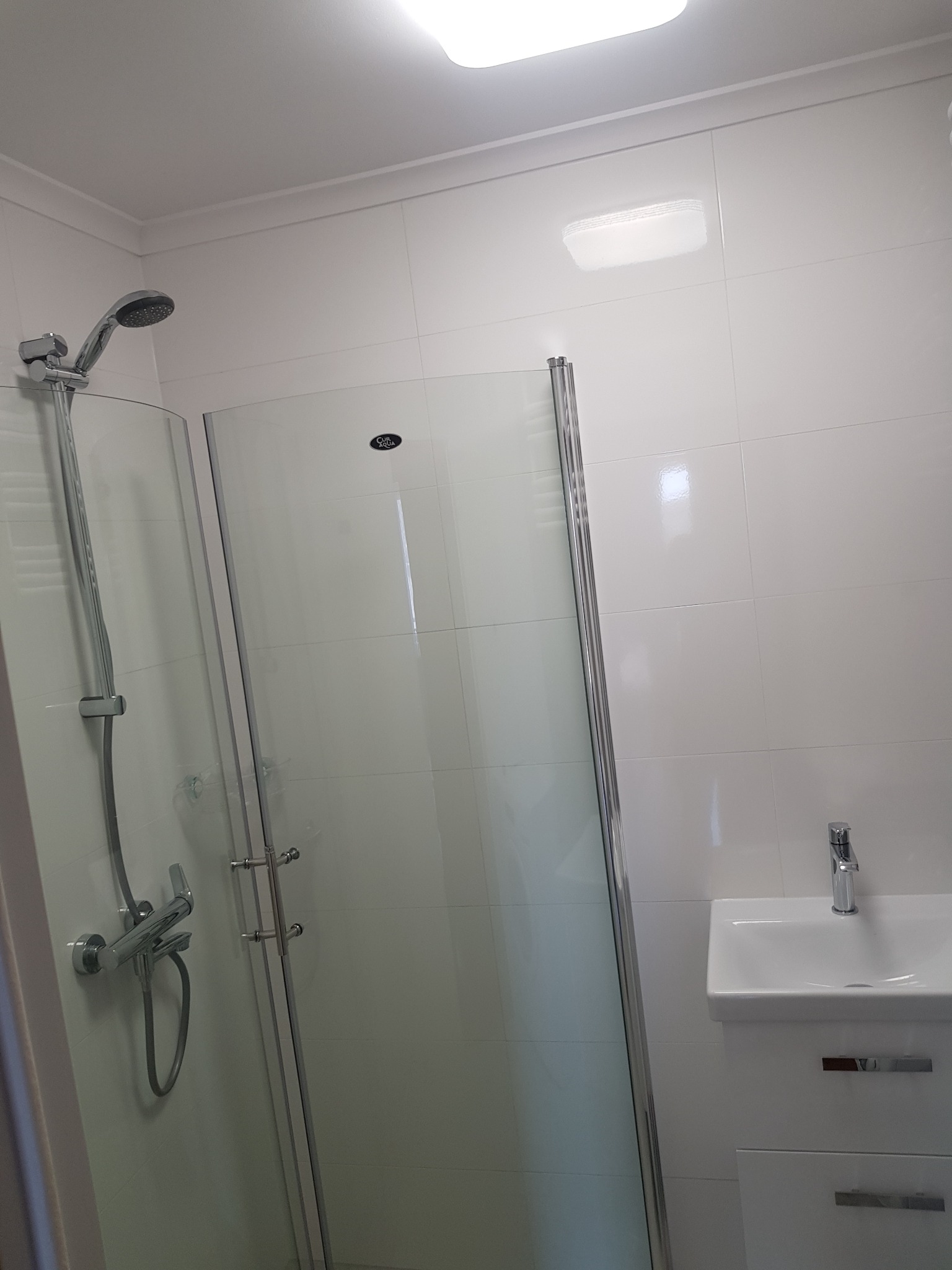 Jasna łazienka z kabiną prysznicową ze szklanymi drzwiami, umywalką nablatową na szafce i chromowaną armaturą na białych płytkach.