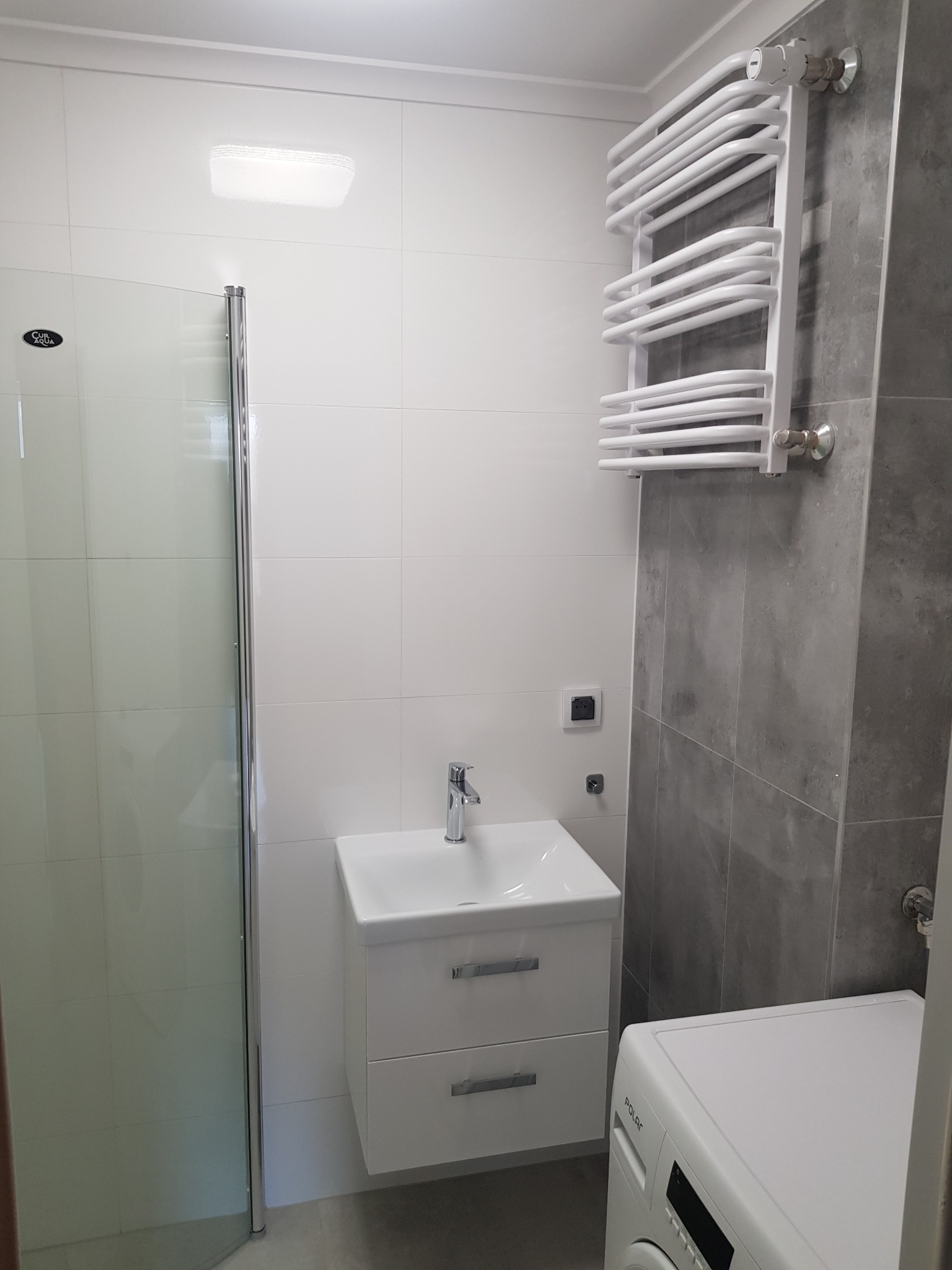 Nowoczesna łazienka z kabiną prysznicową, białą umywalką z szafką, grzejnikiem drabinkowym na szarej ścianie i pralką w rogu.