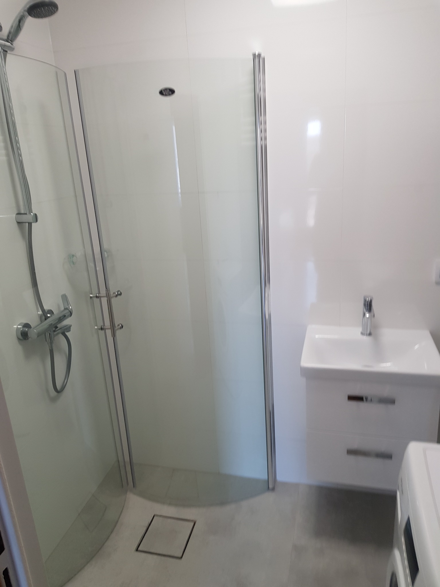 Nowoczesna łazienka z kabiną prysznicową z przezroczystego szkła, białą umywalką z szafką i szarymi płytkami podłogowymi.