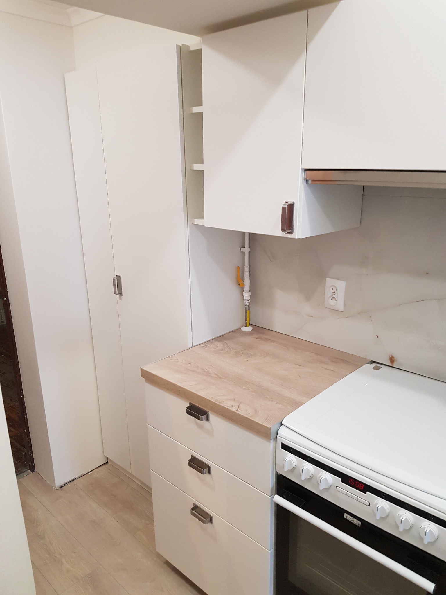 Nowoczesna, jasna kuchnia z białymi szafkami, blatem w kolorze jasnego drewna i białą kuchenką. Widoczny fragment instalacji gazowej. Minimalistyczne uchwyty szafek.