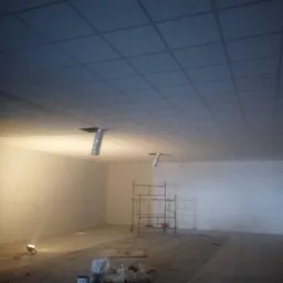 Montaż sufitu podwieszanego z widocznymi elementami wentylacji, rusztowaniem i materiałami budowlanymi w surowym wnętrzu. Oświetlenie punktowe, przygotowanie do prac wykończeniowych.