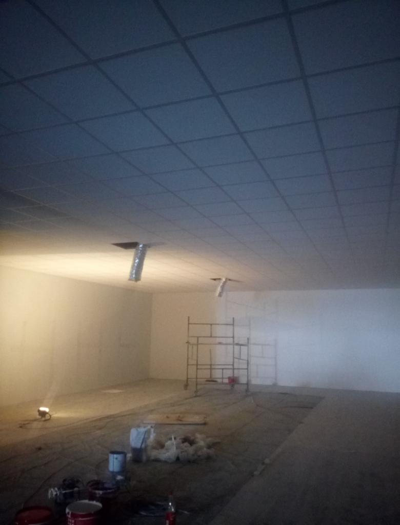 Montaż sufitu podwieszanego z widocznymi elementami wentylacji, rusztowaniem i materiałami budowlanymi w surowym wnętrzu. Oświetlenie punktowe, przygotowanie do prac wykończeniowych.