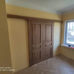 Zamknięte drewniane drzwi przesuwne w stylu rustykalnym z widocznymi, drobnymi uchwytami na tle żółtej ściany i parkietu ułożonego w jodełkę.