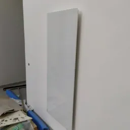 Biały panel grzewczy na ścianie w trakcie montażu, widoczny fragment podłogi i narzędzia w tle.