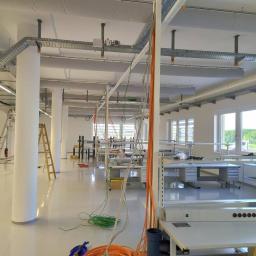 Michał Latosek - Wnętrze jasnego pomieszczenia w trakcie instalacji tras kablowych pod sufitem, widoczne drabiny i zwisające przewody w kolorach pomarańczowym i zielonym.