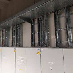Michał Latosek - Rozbudowana instalacja elektryczna w budynku przemysłowym, widoczne szyny montażowe, kable prowadzone w korytach pod sufitem oraz szafy rozdzielcze z symbolami ostrzegawczymi.