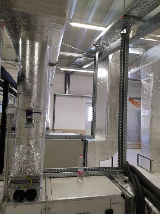 System wentylacji przemysłowej z izolowanymi kanałami, widoczne metalowe koryta kablowe pod sufitem, butelka wody na urządzeniu.