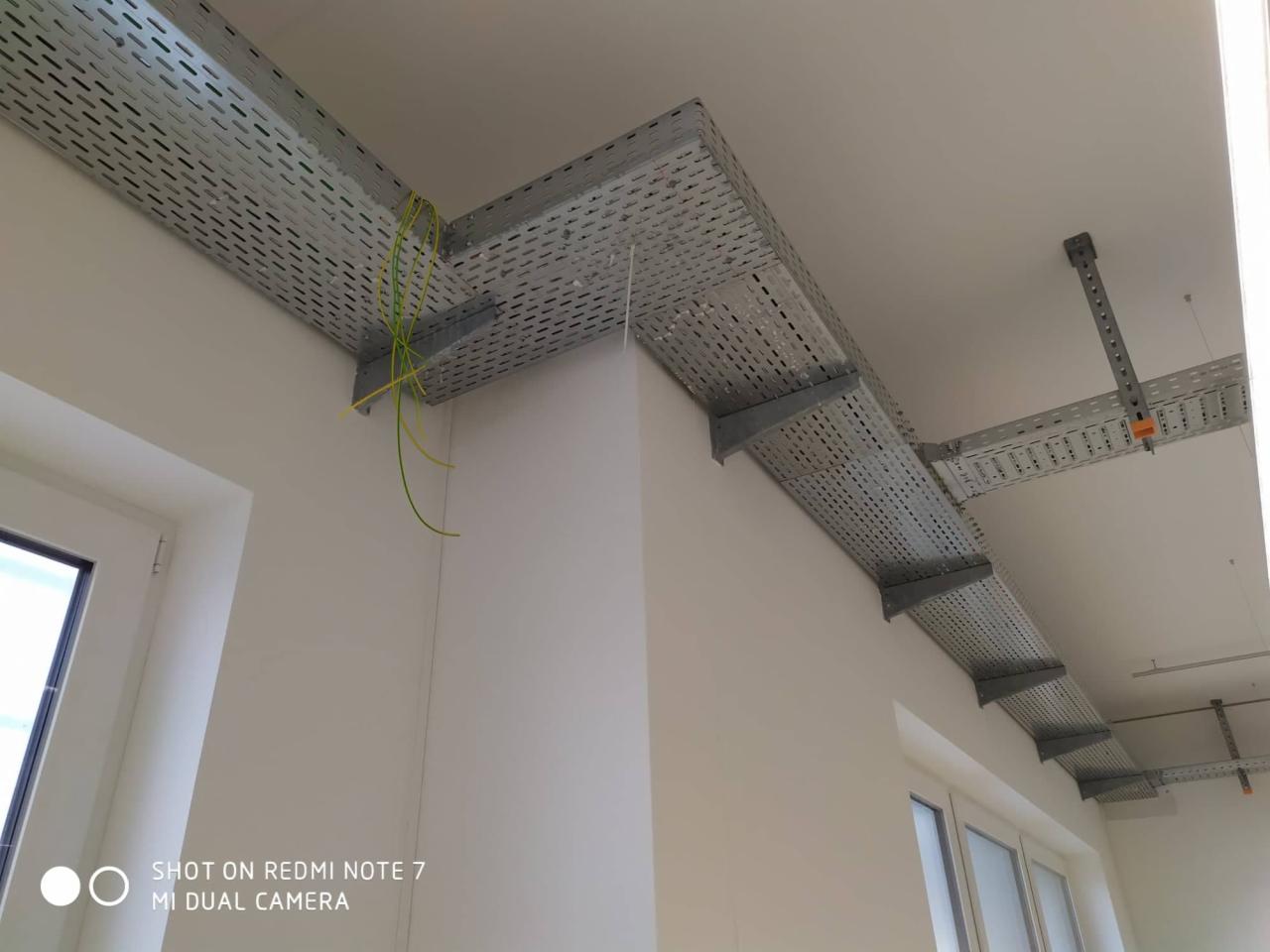 Metalowe korytka kablowe zamocowane pod sufitem w pomieszczeniu, widoczne wystające przewody elektryczne i fragment okna.