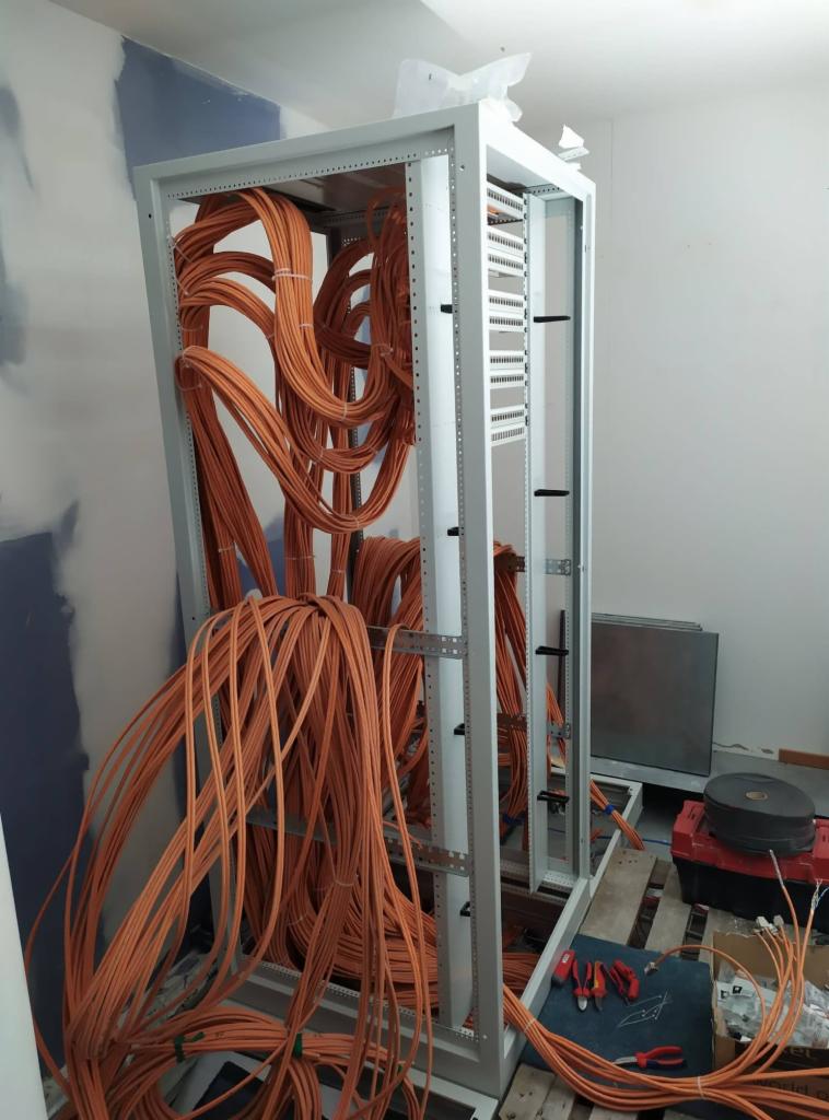 Szafa serwerowa w trakcie instalacji okablowania, widoczne pomarańczowe kable układane wewnątrz ramy, narzędzia leżące na podłodze na palecie.