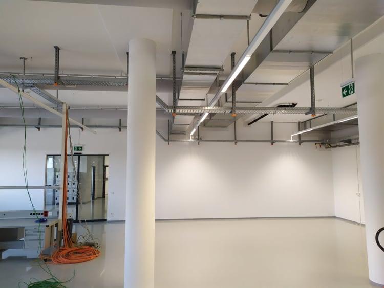 Wnętrze hali produkcyjnej z kolumnami, widoczne koryta kablowe pod sufitem i zwisające przewody elektryczne przy stanowisku pracy.