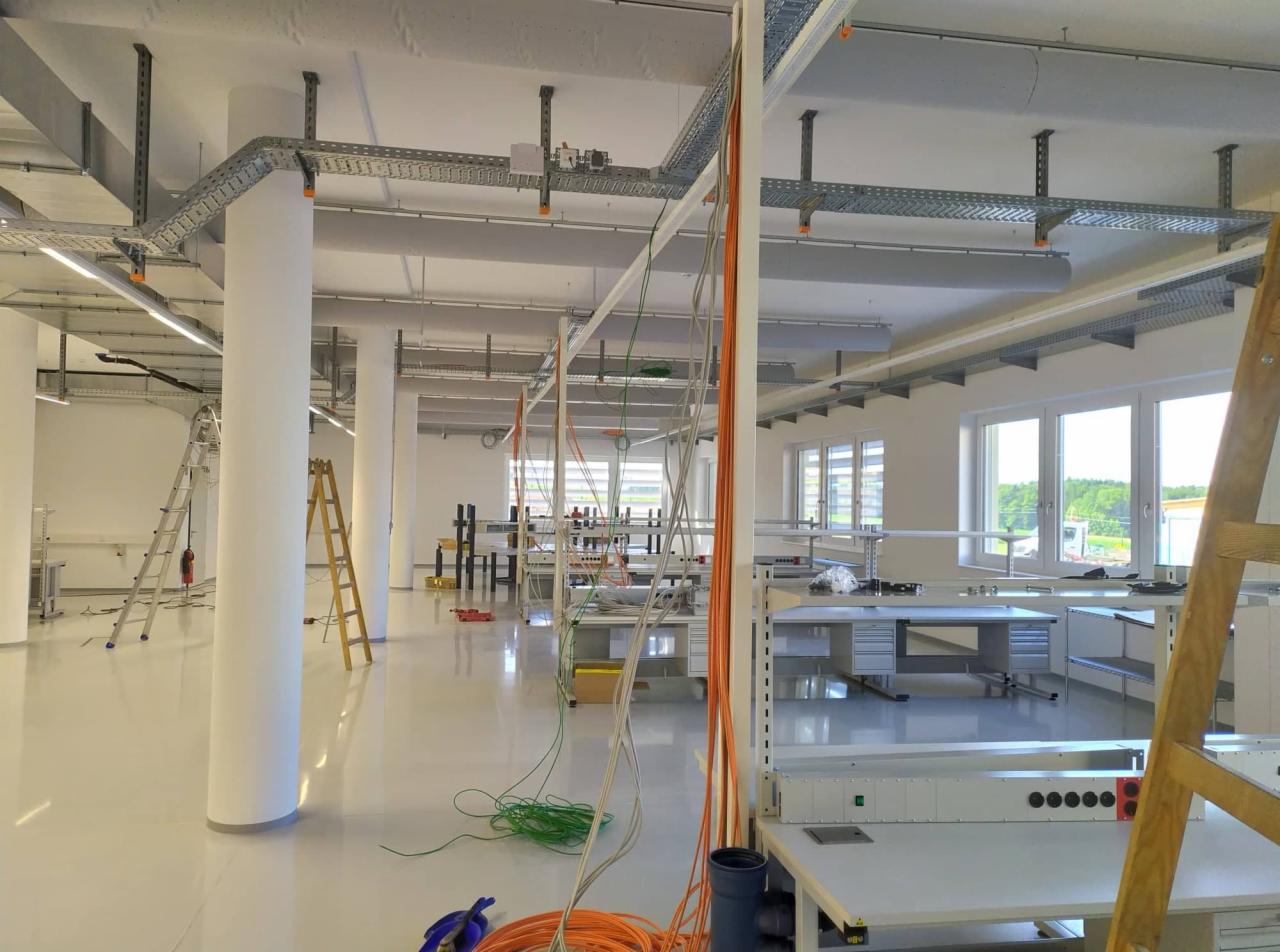 Wnętrze jasnego pomieszczenia w trakcie instalacji tras kablowych pod sufitem, widoczne drabiny i zwisające przewody w kolorach pomarańczowym i zielonym.