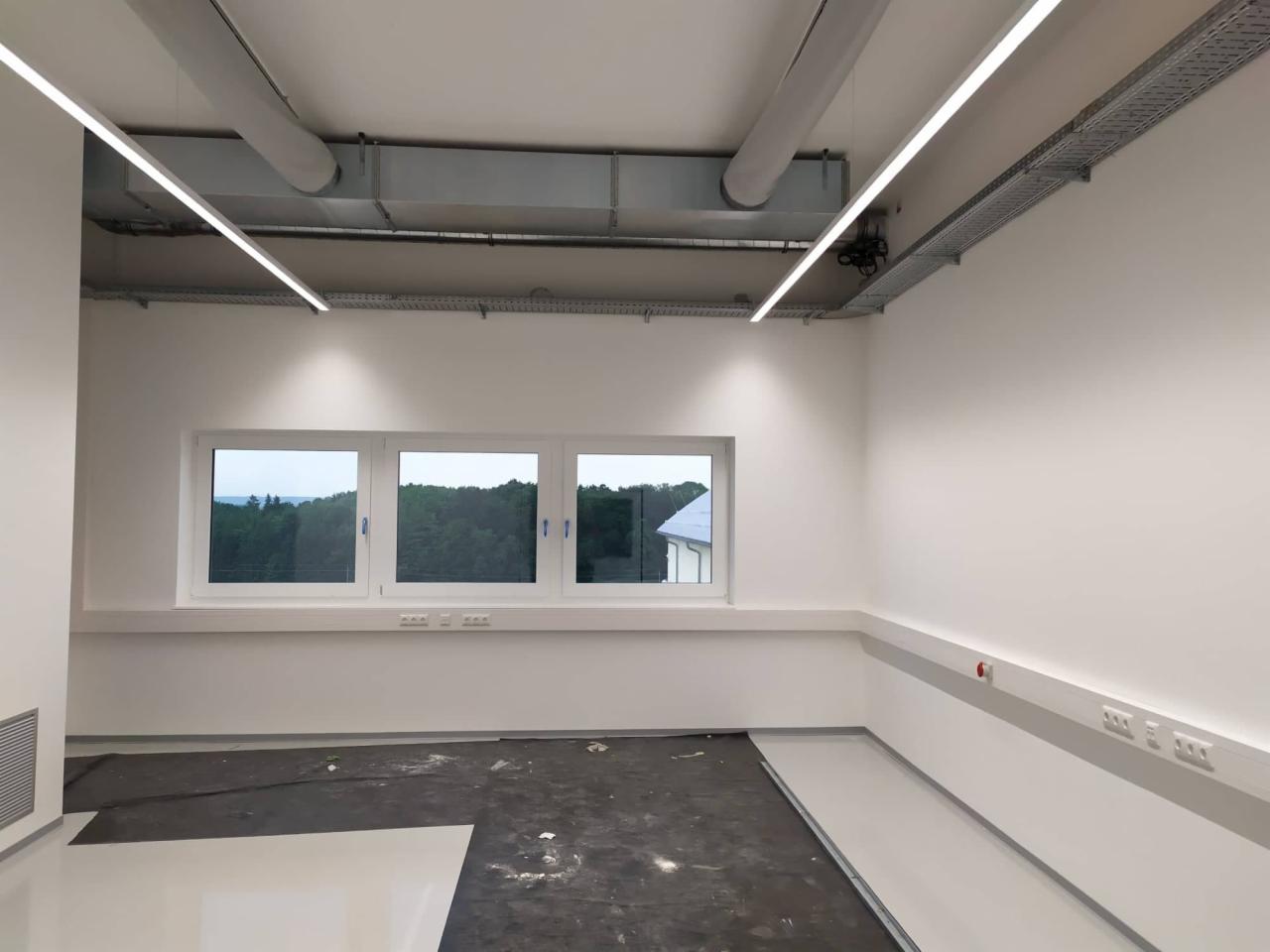 Wnętrze biurowe z nową instalacją oświetleniową: liniowe lampy LED pod sufitem, widoczne koryta kablowe i wentylacja, okna z widokiem na zieleń, częściowo zabezpieczona podłoga w trakcie prac...