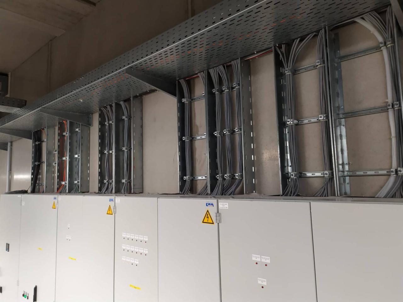 Rozbudowana instalacja elektryczna w budynku przemysłowym, widoczne szyny montażowe, kable prowadzone w korytach pod sufitem oraz szafy rozdzielcze z symbolami ostrzegawczymi.