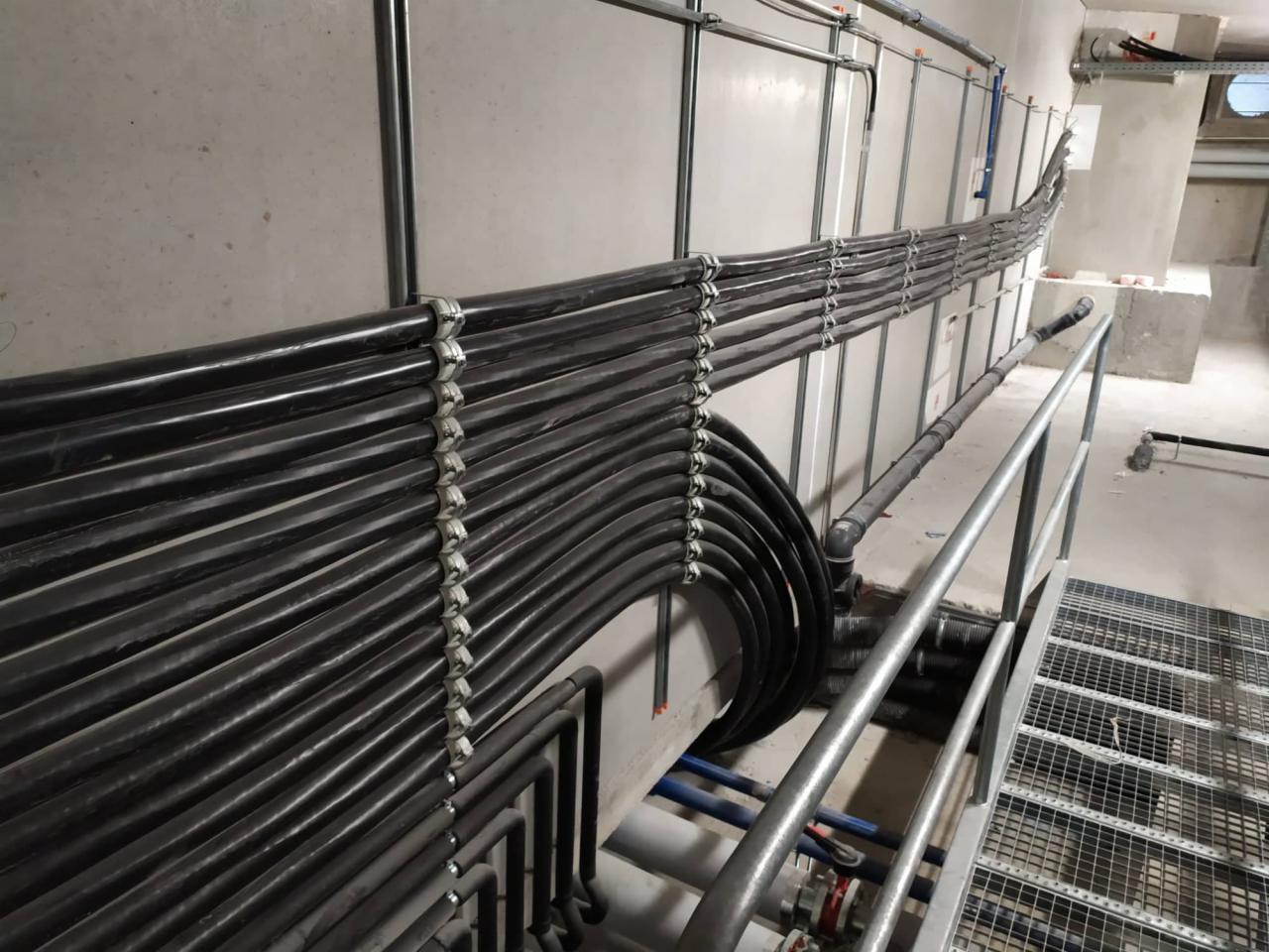 Równolegle ułożone rury w czarnej izolacji, przymocowane do szarej ściany metalowymi uchwytami, widoczne fragmenty instalacji wodnej i metalowa kratka podłogowa.