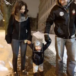 Rodzinne zdjęcie z zimowego spaceru. Kobieta i mężczyzna trzymają za ręce małe dziecko w czapce z uszami. W tle ośnieżone drzewa i budynek.