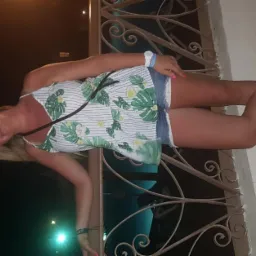 Kobieta w letniej stylizacji, z różowym manicure, pozuje przy ozdobnej balustradzie w wieczornym oświetleniu. Widoczne błyszczące buty i zegarek na ręce.