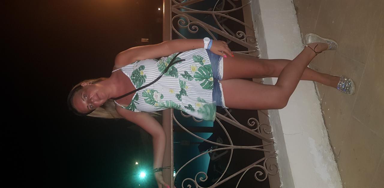 Kobieta w letniej stylizacji, z różowym manicure, pozuje przy ozdobnej balustradzie w wieczornym oświetleniu. Widoczne błyszczące buty i zegarek na ręce.