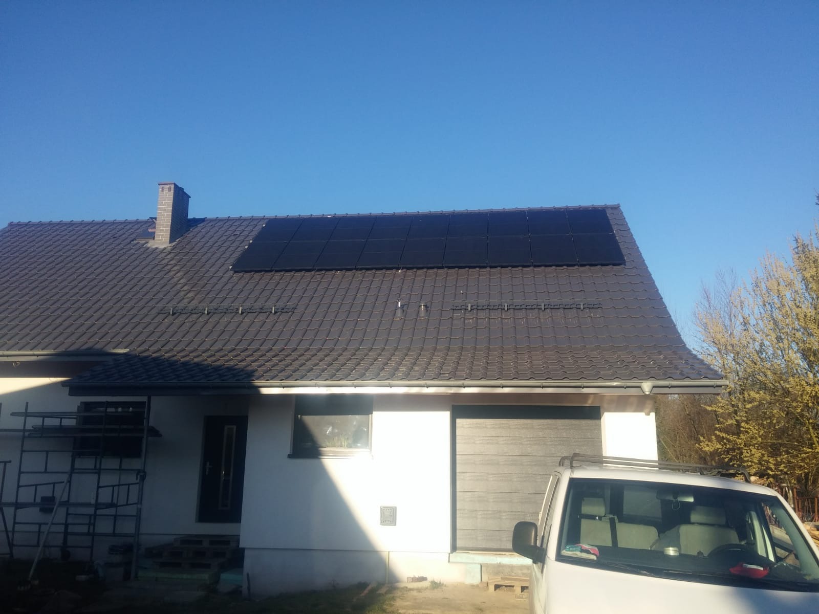 Dom jednorodzinny z ciemną dachówką i panelami fotowoltaicznymi na dachu, widoczny również samochód dostawczy i rusztowanie przy ścianie.