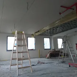Wnętrze w trakcie montażu sufitu z płyt kartonowo-gipsowych, widoczny podnośnik do płyt, drabiny i zabezpieczone folią okna.
