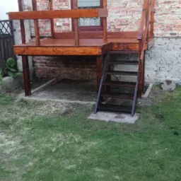 Drewniany taras z balustradą i schodami, przylegający do ceglanego budynku z oknem, widok z poziomu trawnika.