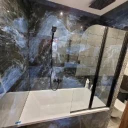 Wykończona łazienka z wanną obłożoną płytkami imitującymi marmur, czarna armatura i szklany parawan nawannowy z czarnymi ramami, widoczny odpływ liniowy.