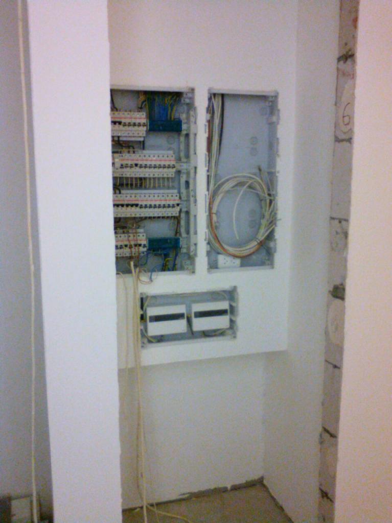 Wnętrze szafki elektrycznej z widocznymi bezpiecznikami, przewodami i listwami zaciskowymi w surowym stanie wykończenia ściany.