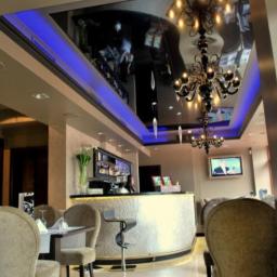 Swiss Company Jmm Sp.Z.O.O - Eleganckie wnętrze lokalu z błyszczącym, czarnym sufitem podświetlonym na niebiesko, ozdobnym żyrandolem, barem z białym frontem i krzesłami w kolorze beżowym.