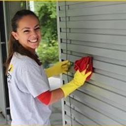 Swiss Company Jmm Sp.Z.O.O - Kobieta w żółtych rękawicach i koszulce firmowej czyści szare żaluzje okienne czerwoną ściereczką, uśmiechając się do kamery.