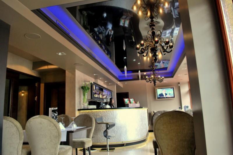 Eleganckie wnętrze lokalu z błyszczącym, czarnym sufitem podświetlonym na niebiesko, ozdobnym żyrandolem, barem z białym frontem i krzesłami w kolorze beżowym.