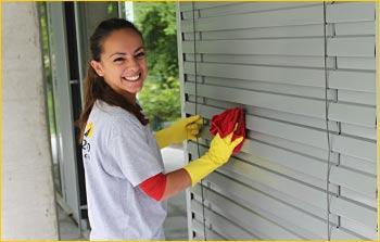 Kobieta w żółtych rękawicach i koszulce firmowej czyści szare żaluzje okienne czerwoną ściereczką, uśmiechając się do kamery.