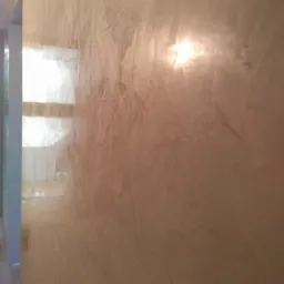 Beżowa ściana z dekoracyjną strukturą, widoczne refleksy światła i fragment okna w tle.