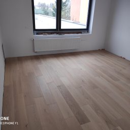 Arrur Musiał - Jasna, dębowa podłoga panelowa w nowym, pustym pokoju z białymi ścianami i grzejnikiem pod oknem; widoczne pomarańczowe kliny dystansowe przy ścianach.