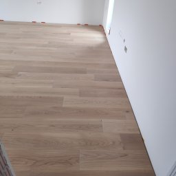 Arrur Musiał - Jasne panele podłogowe ułożone w nowym pomieszczeniu, widoczne pomarańczowe kliny dystansowe przy ścianie, przygotowane puszki elektryczne.