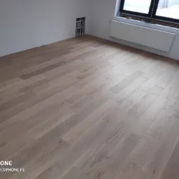 Jasne panele podłogowe ułożone w nowym pomieszczeniu z grzejnikiem pod oknem i instalacją hydrauliczną w ścianie.